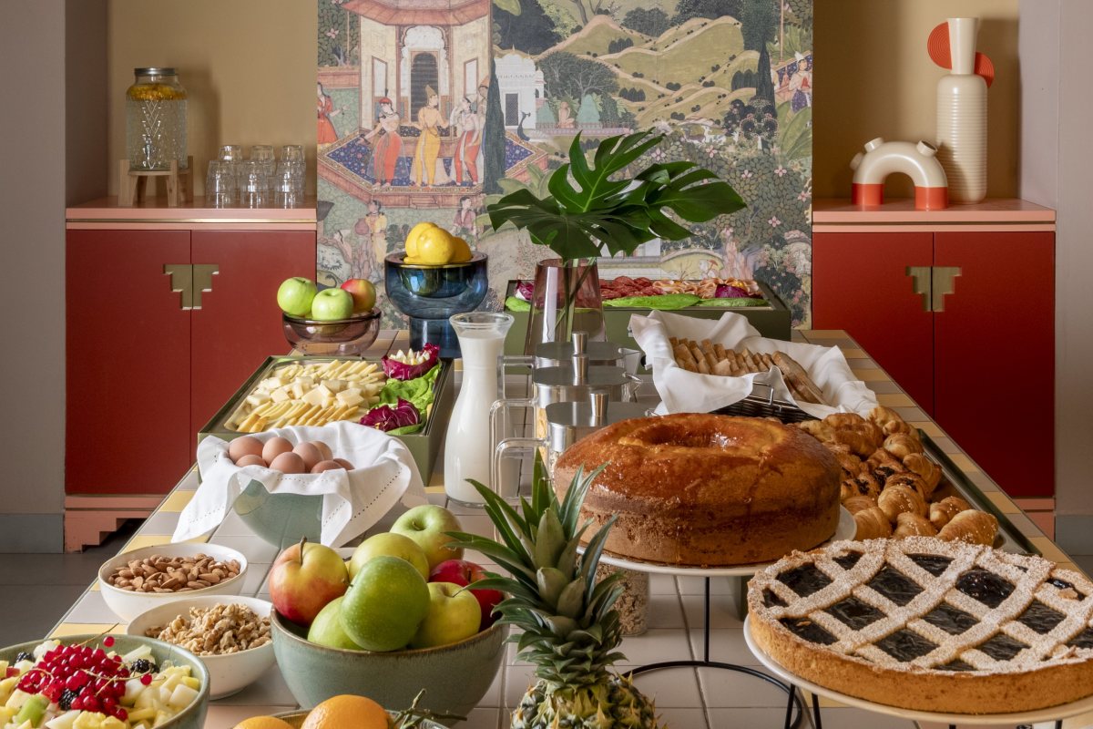 Rich breakfast buffet with tarts, bundt cakes, eggs, cheeses, and fresh fruit, presented on a tiled table in front of a decorated screen - Hotel Milani, BZAR hotels in Rome
