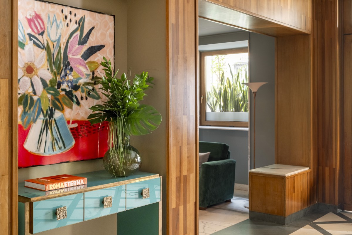 Close-up of a light blue console table with gold knobs, topped with a colorful floral painting and a vase of tropical leaves in a wood-paneled hallway - Hotel Milani, BZAR hotels in Rome